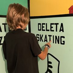 A kid pointing to the Deleta Skating space on the giant game-board wall in Gameland